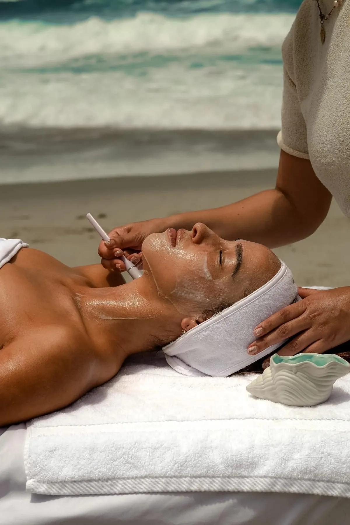 An esthetician brushing a treatment mask onto a client's face, with the ocean behind them